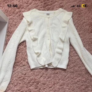 Vit stickad kofta med volang från Lindex - Supersöt vit stickad kofta från Lindex med volangdetaljer längs axlar och framstycke. Koftan har rund hals, knappar framtill och långärmad passform. Perfekt för lager på lager och ger en gullig touch till din outfit.