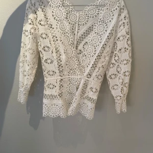 Vit spetsblus från Sandro Paris - Superfin vit blus från Sandro Paris med v-ringad omlottfront och detaljerad spets. Blusen har trekvartsärm och vackra broderade mönster över hela plagget. Perfekt för dig som gillar romantisk och feminin stil med extra detaljer.