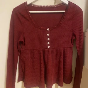 Vinröd ribbad blus med spetskant - Säljer en vinröd blus med ribbad struktur och söt spetskant runt halsringningen. Blusen har lång ärm, rundad hals och fem vita knappar framtill. Modellen är lätt utställd nedtill för en skön passform. Perfekt för dig som gillar romantiska detaljer.