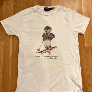 Vit Polo Bear t-shirt från Ralph Lauren - Vit t-shirt från Polo Ralph Lauren med ikoniskt Polo Bear-tryck där björnen åker skidor. Klassisk rund hals, korta ärmar och smal passform (custom slim fit). Tillverkad i mjuk bomull, perfekt för en chill och stilren look.