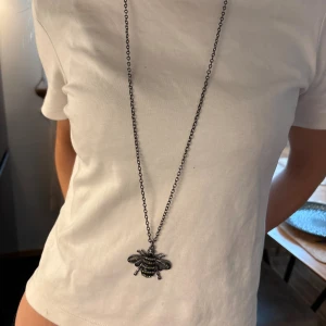 Långt halsband med bi-hänge - Unikt långt halsband i silverfärg med ett stort bi som hänge. Biet har glittriga detaljer och svarta ränder, vilket ger en cool och lekfull look. Kedjan är tunn och stilren, perfekt för att sticka ut med en statement-accessoar.