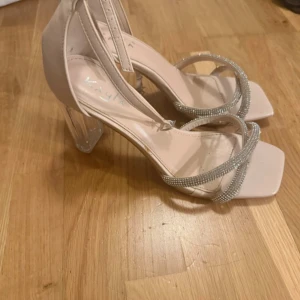 Beiga sandaler med strass och klack - Snygga beiga sandaler från Kaya med transparent hög klack och glittriga strassband över foten. Skorna har fyrkantig tå och justerbar rem runt vristen. Perfekta för dig som vill sticka ut med en elegant och trendig look.