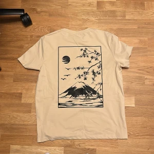 Beige t-shirt med japanskt tryck SHEIN - Beige t-shirt från SHEIN i storlek XXL med ett stort svart tryck på ryggen som föreställer ett japanskt landskap med berg, sol, fåglar och blommande grenar. Klassisk rund hals och korta ärmar. Skön och mjuk känsla, perfekt för en chill stil.