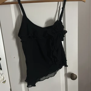 Svart linnetopp med volang och blomdetalj - Snygg svart linnetopp med smala axelband och volangdetaljer. Toppen har en dekorativ blomma i tyg på ena axelbandet och asymmetrisk nederkant. Tunt, lätt material som ger en luftig känsla. Den går snett ner vid sidan av jeansen och är super fin bara att den inte kommer till användning 😅💗 Användt 2 gånger ‼️