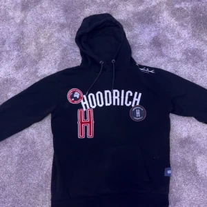 Svart hoodie från Hoodrich med tryck - Svart hoodie från Hoodrich i storlek S med stor vit HOODRICH-text och röda samt svarta patchar på bröstet. Tröjan har huva med snörning och en klassisk känguruficka framtill. Materialet är mjukt och känns som bomull, perfekt för chill dagar.