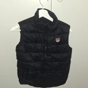 Svart dunväst från GANT - Svart dunväst från GANT med quiltad design och hög krage. Västen har dragkedja framtill och ett broderat GANT-emblem på bröstet. Perfekt för lager-på-lager och har en stilren look som funkar till många outfits.