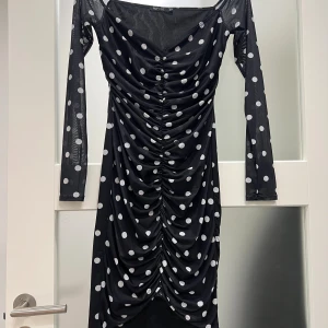 Svart prickig meshklänning - Svart meshklänning i storlek XS med vita prickar, långärmad och med snygg rynkning framtill. Klänningen har en v-ringad hals och är figurnära med stretchigt material som ger en cool och trendig look.