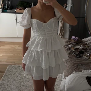 Vit kort klänning med puffärm och volanger - Supersöt vit kort klänning med puffärmar och flera lager volanger i kjolen. Klänningen har markerad byst med sömmar och smock i ryggen, vilket ger en snygg passform. Materialet är strukturerat och lätt, perfekt för varma dagar. Dragkedja bak för enkel påklädning.