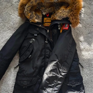 Svart Parajumpers parkajacka med päls - Säljer en svart parkajacka från Parajumpers med stor fluffig päls runt huvan och snygga detaljer som gul rem och flera fickor. Jackan är quiltad på insidan och har dragkedja framtill. Perfekt för kalla dagar när du vill hålla stilen.