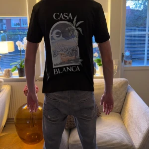 Svart Casa Blanca t-shirt med tryck - Svart t-shirt från Casa Blanca med coolt grafiskt tryck på ryggen och mindre tryck på bröstet. T-shirten har korta ärmar och en klassisk rund hals. Materialet känns mjukt och skönt, perfekt för dig som gillar streetwear med unik design.