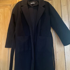 Svart kappa från Pieces med bälte - Svart lång kappa från Pieces med klassisk krage och två stora fickor framtill. Jackan har ett snyggt bälte i midjan för att kunna knyta och justera passformen. Perfekt för dig som gillar stilrena och enkla plagg med lite edge. Lite för liten på mig nu. Inga defekter! Behöver bara håras av med en roller. Obs den är inte fodrad inuti!