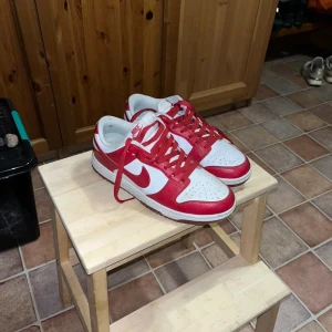 Nike Dunk Low röd/vit sneakers - Nike Dunk Low sneakers i rött och vitt med klassisk Swoosh-logga på sidorna. Skorna har röd snörning, perforerad tåbox och vit meshfoder. Yttermaterialet är skinn och sulan är platt. Perfekt för dig som gillar streetstyle och vill sticka ut.