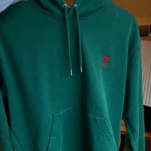 Grön hoodie från Ami Paris med hjärtdetalj - Snygg grön hoodie från Ami Paris med röd broderad hjärtdetalj på bröstet. Tröjan har huva med snörning, stor magficka och ribbade muddar. Perfekt för dig som gillar stilrena och coola plagg med en twist. Nypris runt 2000! Kan skicka fler bilder om frågan finns. Öppen för prisförslag!