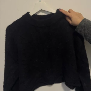 Svart fluffig stickad tröja från H&M - Mysig svart stickad tröja med fluffig yta och långärmad modell. Tröjan har en enkel rund halsringning och är lite croppad i passformen, perfekt för att ge en chill vibe till din outfit. Passar dig som gillar basic men ändå vill ha något extra mjukt och snygg.