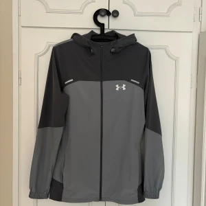 Grå vindjacka från Under Armour  - Snygg tvåfärgad grå vindjacka från Under Armour med huva och hel dragkedja. Jackan har logga på bröstet och tryckt text längs ryggen. Perfekt för träning eller när du vill ha en sportig look. Tillverkad i lätt syntetmaterial och har elastiska muddar. Använd fåtal gånger och my let bra skick
