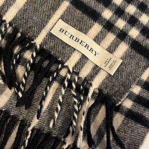 Rutig halsduk i kashmir från Burberry - Exklusiv halsduk från Burberry i klassiskt rutigt mönster med svarta, gråa och beige toner. Tillverkad i 100% kashmir och har snygga tvinnade fransar i ändarna. Perfekt accessoar för att lyfta din outfit med en tidlös brittisk känsla.