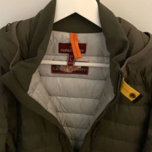 Olivgrön dunjacka från Parajumpers - Säljer en olivgrön dunjacka från Parajumpers med huva och ikonisk gul detalj vid halsen. Jackan har quiltad design, dragkedja framtill och patch med logga på ärmen. Perfekt för dig som vill ha en lätt och snygg jacka med streetkänsla.