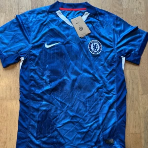 Chelsea FC Palmer 10 matchtröja Nike - Chelsea FCs matchtröja med Palmer 10-tryck på ryggen. Tröjan är blå med subtila mönster, vita detaljer och röda inslag på sidorna. Broderat klubbmärke på bröstet och Nike-logga. Tillverkad i lätt och ventilerande polyester.