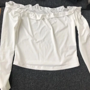 Vit offshoulder topp med volang - Supersöt vit offshoulder topp med långa ärmar och volangdetalj längs halsringningen. Toppen har en liten rosett framtill och är gjord i ett mjukt, stretchigt material. Aldrig använd💕