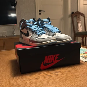 Nike Air Jordan 1 High OG University Blue - Nike Air Jordan 1 High OG i färgen University Blue med vit och grå ovandel i skinn, svarta swoosh-detaljer och ljusblå snörning samt sula. Klassisk Jordan Wings-logga på sidan och ikonisk design som sticker ut. Perfekt för dig som vill ha en snygg och sportig look.