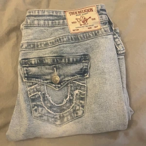 True Religion Joey ljusblå flare jeans - Säljer ett par True Religion Joey jeans i ljusblå tvätt med låg midja och flare-ben. Klassisk femficksmodell med snygga detaljer på bakfickan och silverfärgad knapp framtill. Materialet är jeans och passformen är loose med bootcut-stil. Passar mig som har M 🌸