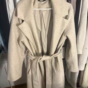 Beige kappa med bälte och bred krage - Snygg beige kappa med bred krage och tillhörande bälte i midjan. Jackan har en klassisk, rak passform och är helfodrad. Perfekt för dig som gillar stilrena och neutrala färger. Materialet känns mjukt och har en ull-liknande struktur. Passar dig som vill ha en tidlös look.
