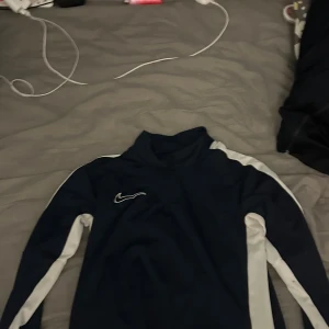 Marinblå Nike långärmad träningströja - Säljer en marinblå långärmad träningströja från Nike med vita ränder längs ärmarna och axlarna. Tröjan har en hög krage och är gjord i ett mjukt syntetmaterial som andas bra. Perfekt för sport eller chill dagar. Snygg och sportig design med Nike-logga på bröstet.