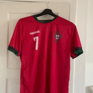 Portugal Ronaldo #7 fotbollströja - Säljer en röd Portugal fotbollströja med nummer 7 och Ronaldo på ryggen. Tröjan har gröna och svarta detaljer vid krage och ärmslut, samt landslagets emblem på bröstet. Tillverkad i lätt och ventilerande material, perfekt för match eller träning.