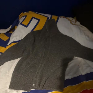 Grå ribbad kofta med dragkedja - Säljer en grå ribbad kofta med hel dragkedja framtill och hög krage. Tröjan har långärmad design och är stickad i ett mjukt material som passar perfekt när det är lite kyligt ute. Enkel och stilren, funkar till många olika looks.