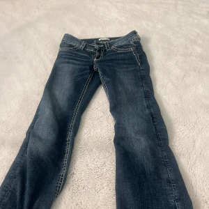 Blå bootcut jeans från Gina Tricot - Snygga blå jeans från Gina Tricot i storlek 152 med bootcut passform. Jeansen har dekorativa broderier i beige och grått på bakfickorna, kontrastsömmar och klassisk femficksdesign. Perfekta för dig som gillar detaljer och en cool stil.