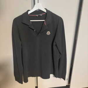 Mörkgrå pikétröja från Moncler - Snygg mörkgrå långärmad pikétröja från Moncler med klassisk krage och två knappar framtill. Tröjan har Moncler-logga på bröstet och diskreta rödvita detaljer på insidan av kragen. Perfekt för dig som gillar stilrena och exklusiva märken.