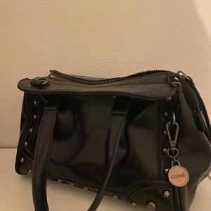 Svart handväska från Conmé med nitar - Snygg svart handväska från Conmé i skinnimitation med silverfärgade nitar längs sidorna. Väskan har dubbla handtag, dragkedja upptill och en rund Conmé-tag i metall. Som ny, använd 2 gånger. Nypris 899kr 