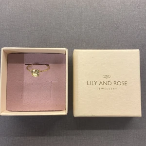 Ring  Lily and Rose - Snygg guldfärgad ring från Lily and Rose med en stor, rund ljus sten i mitten. Ringen har en hamrad yta som ger en cool och unik look. Kommer i original ask, perfekt för dig som gillar stilrena och trendiga smycken.