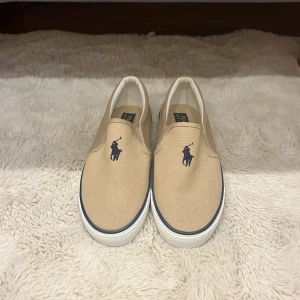 Beige slip-on sneakers Polo Ralph Lauren - Snygga slip-on sneakers från Polo Ralph Lauren i beige canvas med klassisk mörkblå broderad logga framtill. Skorna har vit och brun gummisula med Polo-tryck baktill och en rund tå. Perfekt för dig som gillar stilren och enkel design.
