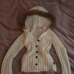 Snygg vintage kofta - Vit stickad kofta med mysig huva kantad av fuskpäls. Koftan har kabelstickat mönster, stora svarta knappar framtill och lång ärm. Perfekt för kyliga dagar när du vill vara både varm och snygg.