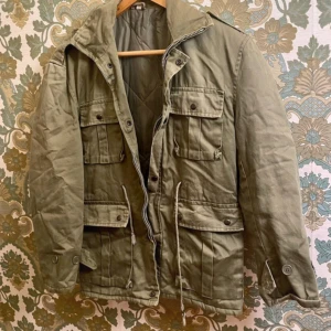 Olivgrön jacka - Parkajacka i olivgrönt med fyra stora fickor framtill, snörning i midjan och klassisk krage. Jackan har långa ärmar med knappdetaljer och dragkedja framtill. Materialet är slitstark bomull med quiltat foder för extra värme.