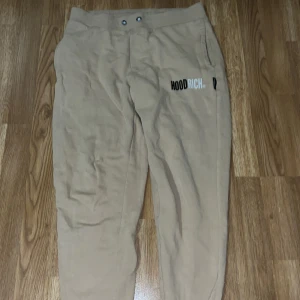 Beige mjukisbyxor från Hoodrich - Snygga beige mjukisbyxor från Hoodrich med tryckt logga på ena benet. Byxorna har normal passform, midwaist och är tillverkade i mjukt bomullsmaterial. Perfekta för chill eller streetwear och har praktiska fickor samt knappdetaljer i midjan.