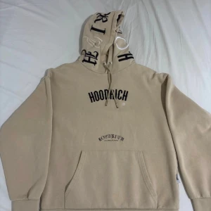 Beige hoodie från Hoodrich med tryck - Säljer en beige hoodie från Hoodrich med svart tryck på bröstet och broderade bokstäver på huvan. Tröjan har känguruficka, snörning i huvan och är gjord i mjuk bomullsmix. Perfekt för chill dagar och streetwear vibes.