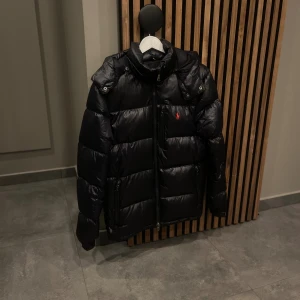 Svart pufferjacka från Polo Ralph Lauren - Svart pufferjacka från Polo Ralph Lauren med huva och dragkedja framtill. Jackan har ett rött Polo-logo broderat på bröstet, flera fickor med dragkedja och hög krage. Perfekt för kalla dagar och har en snygg, modern look.