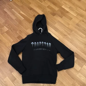 Svart Trapstar hoodie med silvertryck - Svart hoodie från Trapstar i storlek M med fet silverfärgat Trapstar-tryck på bröstet och texten 'It's a secret' under. Hoodien har snörning i huvan och en stor magficka framtill. Perfekt för dig som gillar streetwear och vill sticka ut.