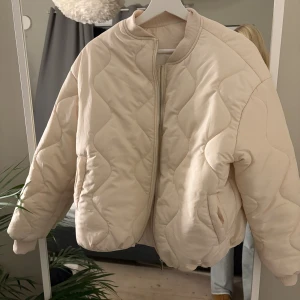 Beige pufferjacka - Säljer en beige pufferjacka med dragkedja framtill. Jackan har rundad krage, långa ärmar och en loose passform. Perfekt för att hålla värmen under kyliga dagar.