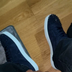 Mörkblå sneakers med nitar - Säljer ett par mörkblå sneakers med vit platt sula och coola nitar på tån. Skorna har snörning och är tillverkade i mocka för en lyxig känsla. Perfekt för dig som vill sticka ut med din stil. DOM ÄR ÄKTA 