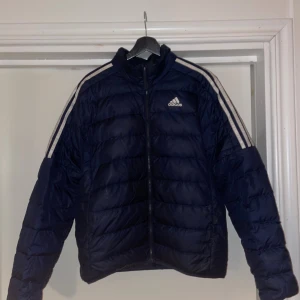 Mörkblå pufferjacka från Adidas - Snygg mörkblå pufferjacka med vita ränder längs ärmarna och Adidas-logga på bröstet. Jackan har dragkedja framtill och quiltad design som ger en sportig vibe. Perfekt för kalla dagar och har en klassisk streetstil. Passar bra för både M och L