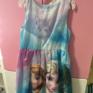 Frozen klänning med rosett - Säljer en söt Frozen-klänning med motiv av Elsa och Anna till barn. i pastelliga blå, rosa och lila toner. Klänningen är ärmlös, har en rund hals och en stor ljusblå rosett baktill. Materialet är glansigt och lätt, perfekt för dig som älskar Frost.