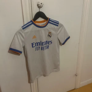 Real Madrid vit matchtröja Adidas - Säljer en officiell Real Madrid fotbollströja från Adidas i vit polyester med blå och orange detaljer. Tröjan har korta ärmar, broderat klubbmärke och tryckt sponsorlogga på bröstet. Klassiska blå Adidas-ränder på axlarna och kontrastfärgade muddar.