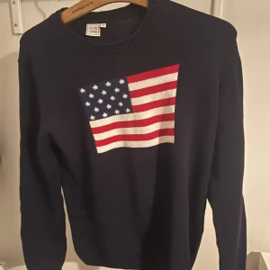 Mörkblå stickad tröja med USA-flagga - Snygg mörkblå stickad tröja från Club XPRS i storlek M. På bröstet finns en stor USA-flagga i rött, vitt och blått. Tröjan har rund hals och långa ärmar, Hör av er vid frågor priset är diskuterbart.