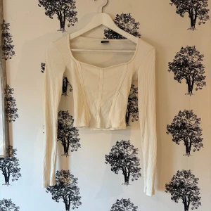 Vit croppad långärmad topp Gina Tricot - Säljer en vit croppad topp från Gina Tricot i ribbad bomull. Toppen har lång ärm, fyrkantig urringning och markerade sömmar framtill som ger en korsettkänsla. Perfekt att styla med jeans eller kjol för en clean look.