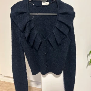 Mörkblå kofta med volang från JDY - Supersnygg mörkblå kofta från JDY med stora volanger längs v-ringningen och knappar framtill. Mjuk stickad känsla och långärmad modell som ger en cool och trendig look. Perfekt för dig som gillar statement-detaljer och vill sticka ut.