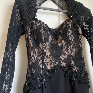 Elegant spetsklänning med pärlor  - Svart långklänning med transparent spets upptill och dekorativa pärlor. Klänningen har långärmade spetsärmar, v-ringad rygg och blommigt spetsmönster. Nedre delen är slät och enfärgad svart, medan överdelen har nude-färgad underklänning. Oanvänd med prislapp kvar
