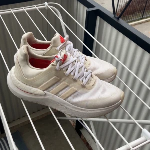 Vita Adidas sneakers med röda detaljer - Snygga vita Adidas sneakers med klassiska tre ränder på sidan och röda detaljer på plösen och hälen. Skorna har en chunky sula med boost-dämpning och ovandel i mesh och syntet. Perfekta för dig som gillar sportig stil och vill ha bekväma skor till vardagen.